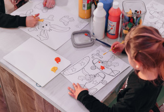 Children painting coloring sheets at a table filled with paints and art supplies in a glamping setting.