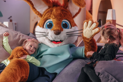Two young girls enjoy time with a large rabbit mascot at a glamping accommodation, smiling happily together.