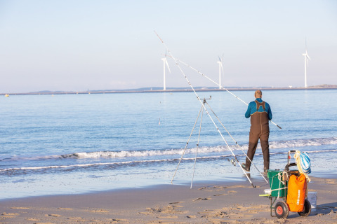 Man fishing on a sandy beach with fishing rods and gear, wind turbines in the background at sea.
