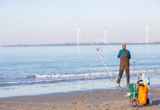 Man fishing on a sandy beach with fishing rods and gear, wind turbines in the background at sea.