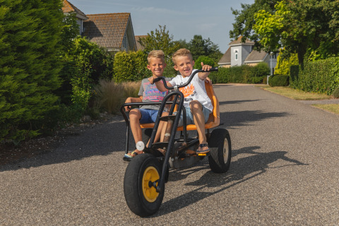 Two smiling children ride a tricycle on a road at a glamping accommodation on a sunny summer day.