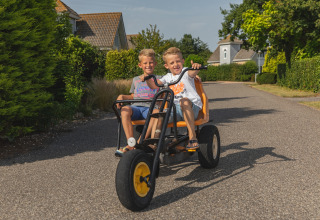 Two smiling children ride a tricycle on a road at a glamping accommodation on a sunny summer day.