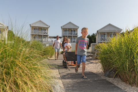 Family with children and wagon arrives at modern glamping cabins surrounded by greenery on a sunny day.