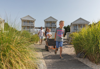 Family with children and wagon arrives at modern glamping cabins surrounded by greenery on a sunny day.