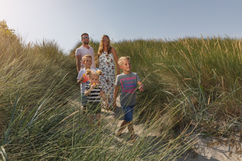 Familie med tre børn går gennem græsset på en sti ved stranden, solskin og afslappet glampingstemning.