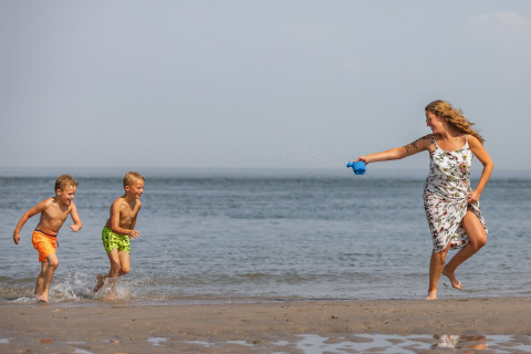 En kvinde i sommerkjole sprøjter vand på to børn ved stranden ved Noordzee Résidence De Banjaard, Holland.