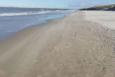 A peaceful sandy beach with distant wind turbines and a dam, viewed from a coastal glamping site.
