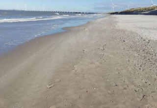 A peaceful sandy beach with distant wind turbines and a dam, viewed from a coastal glamping site.
