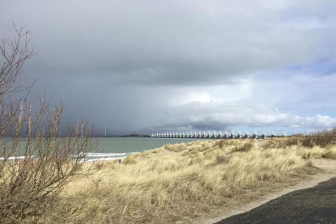 Vue depuis un hébergement glamping sur des dunes, la mer, un ciel nuageux et une digue au loin.