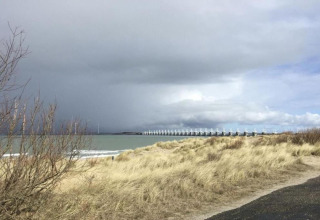 View from a glamping site with sand dunes, sea, cloudy sky, and a long storm surge barrier in the distance.