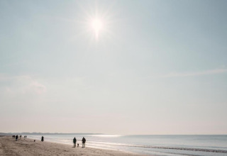 Sunny beach day with people walking by the shoreline, near a glamping accommodation in the distance.