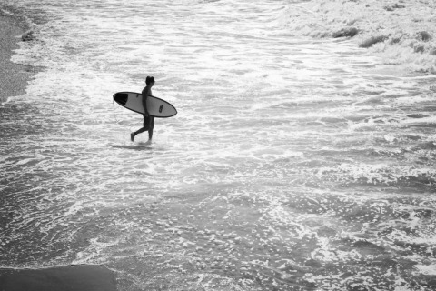Une personne avec une planche de surf marche dans les vagues près du rivage en hébergement glamping.