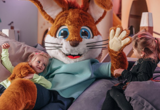 Two children enjoy relaxing with a large bunny mascot in a cozy glamping accommodation setting.