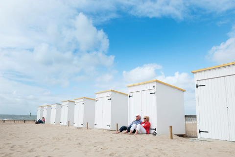 Una pareja sentada frente a cabañas blancas de glamping en la playa bajo un cielo azul parcialmente nublado.