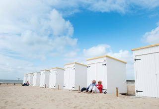 Una pareja sentada frente a cabañas blancas de glamping en la playa bajo un cielo azul parcialmente nublado.