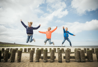 Tre glade børn hopper og leger på stranden foran glamping-indkvartering under en blå himmel.