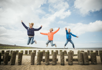 Trois enfants heureux sautent et jouent sur la plage près d’un hébergement glamping sous ciel bleu.