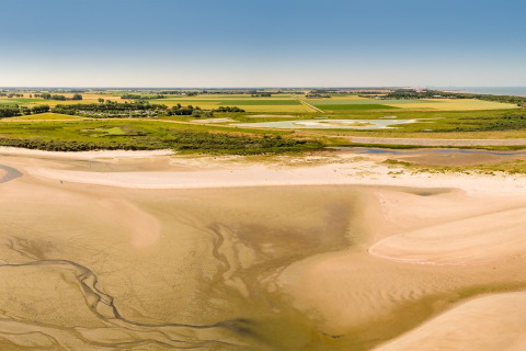 Landskab med sandstrand, grønne marker og glamping-område under en klar blå himmel, sommerstemning.