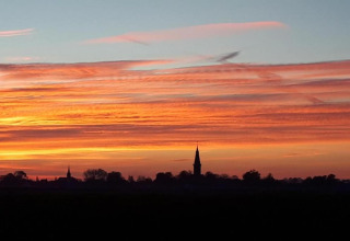 Solnedgang over en glamping-plads med silhuet af kirketårn og farverige skyer i horisonten.