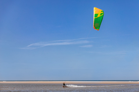En person kitesurferer ved stranden under en blå himmel, fotograferet fra et glampingophold ved kysten.