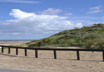 Vista desde un alojamiento glamping con playa de arena, dunas verdes y cielo azul con nubes blancas.