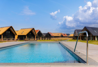 Outdoor swimming pool with modern lodges at Resort Nieuwvliet-Bad 13, Netherlands, under a clear sky.