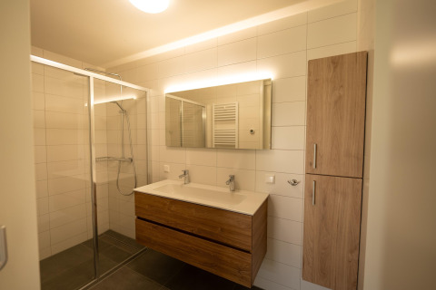 Modernes Bad mit Glasdusche, Doppelwaschbecken, beleuchtetem Spiegel und Holzschränken in Zeebad 14, Niederlande.