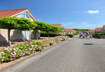 Børn cykler på en vej mellem lodge-hytter med røde tage, træer og blomstrende buske under blå himmel.