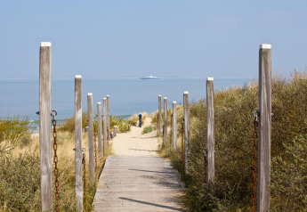 A wooden walkway leads through dunes and bushes toward the beach with sea view and distant boat.