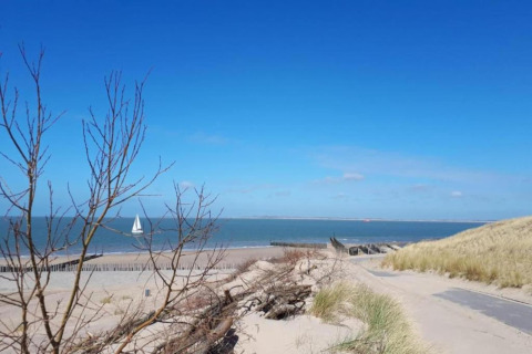 Vista da un lodge su dune di sabbia, spiaggia, mare e una barca a vela sotto cielo azzurro.