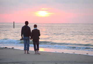 Two people walk barefoot on the beach at sunset, enjoying the tranquil sea and the calming view ahead.