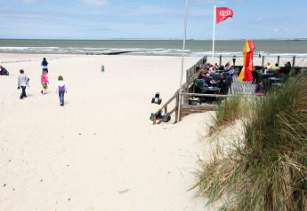 Folk nyder en dag på stranden med caféplads og havudsigt, børn leger i sandet og nogle går en tur.