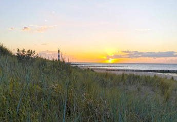 Sunset over the beach with a lighthouse and dunes, seen from a lodge on the coast with tall grasses.