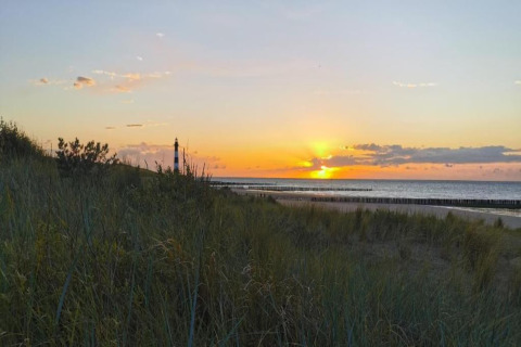 Solnedgang over stranden og en fyrtårn set fra en lodge, omgivet af klitter og græs ved kysten.