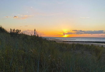 Sunset over the beach with a lighthouse and dunes, seen from a lodge on the coast with tall grasses.