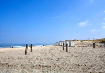 Strandudsigt med hvide hytter, pæle i sandet og folk i det fjerne under en blå himmel ved en lodge.
