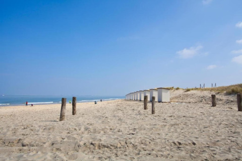 Sandy beach view with white cabins, wooden posts and a few people under blue sky at a seaside lodge.