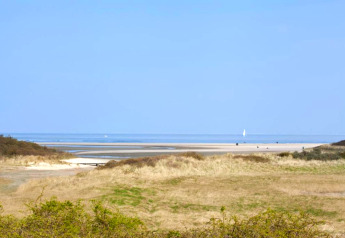 Udsigt fra en lodge over klitter, strand og hav under en klar blå himmel, med en sejlbåd i det fjerne.