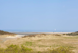 Udsigt fra en lodge over klitter, strand og hav under en klar blå himmel, med en sejlbåd i det fjerne.