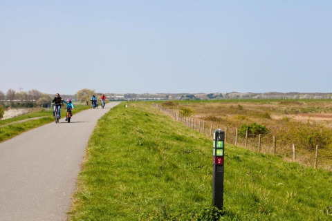 Ciclistas recorren un sendero asfaltado junto a campos verdes y una señal, bajo cielo despejado, cerca de un albergue.