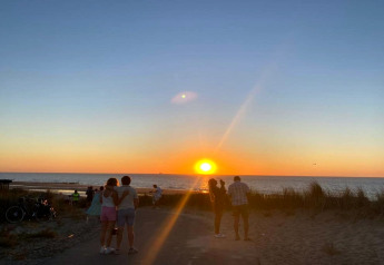 Personer nyder en smuk solnedgang ved havet, omgivet af strand og klitter ved et sommerhus.