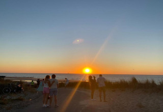 Personer nyder en smuk solnedgang ved havet, omgivet af strand og klitter ved et sommerhus.