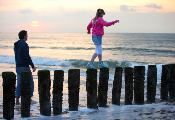 En pige balancerer på træpæle ved stranden ved solnedgang, mens en mand ser til ved hendes side.