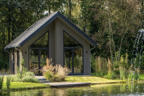 Moderne lodge ved søen i Holiday Park Latour 4, omgivet af grøn vegetation og natur, træhus med glasfacade.