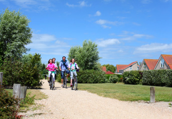 En familie cykler sammen ad en grussti ved ferieboliger og grønne buske under en blå himmel.