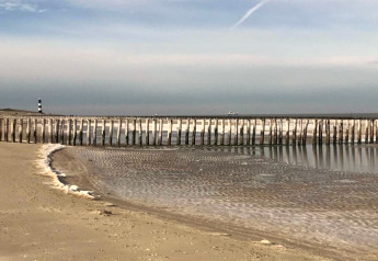 Strandbillede med stolper langs kysten, lavvande og et fyrtårn i det fjerne under en klar himmel.