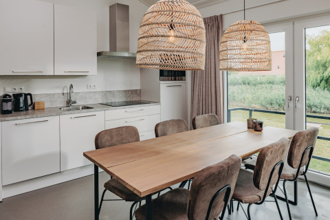 Modern kitchen and dining area with wooden table, six chairs, large windows and wicker lamps at Holiday Park Zeebad 12.