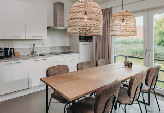 Modern kitchen and dining area with wooden table, six chairs, large windows and wicker lamps at Holiday Park Zeebad 12.