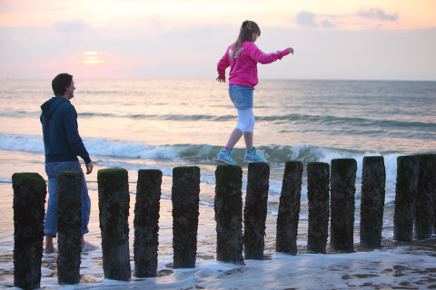A girl balances on wooden posts by the sea at sunset, watched by a man at Holiday Park Zeebad 12 in the Netherlands.