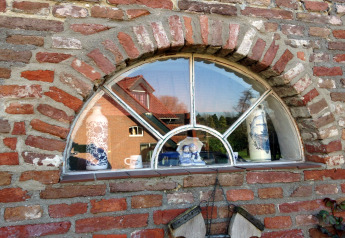 Halvrundt vindue med porcelænsfigurer i vindueskarmen og spejling af et rødt hus i baggrunden.
